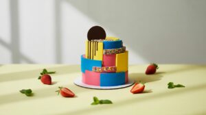 A vibrant, two-tiered cake with geometric blue, pink, and yellow frosting, topped with a chocolate disc and rainbow sprinkles. Strawberries and mint leaves are arranged around the cake on a light yellow surface.